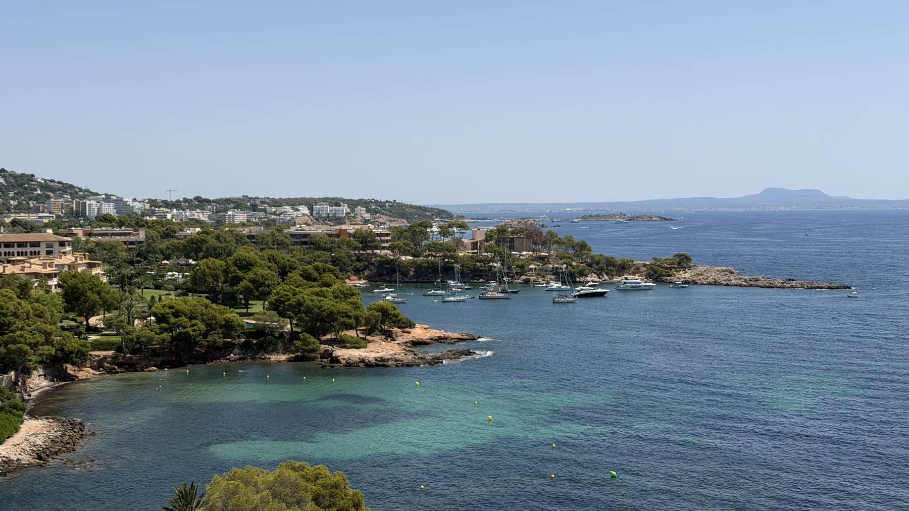 A turquoise Mallorcan cove seen from the headland above, with boats at anchor and pines along the shore